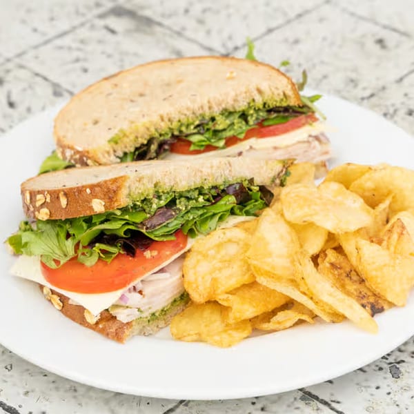 A turkey sandwich with pesto, lettuce, tomato, and cheese, served with a side of potato chips.