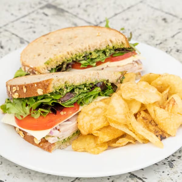 A turkey and pesto sandwich on whole grain bread with lettuce, tomato, and cheese, served with chips.