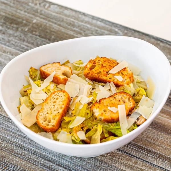 A Caesar salad in a white bowl, topped with large croutons and shaved Parmesan cheese.