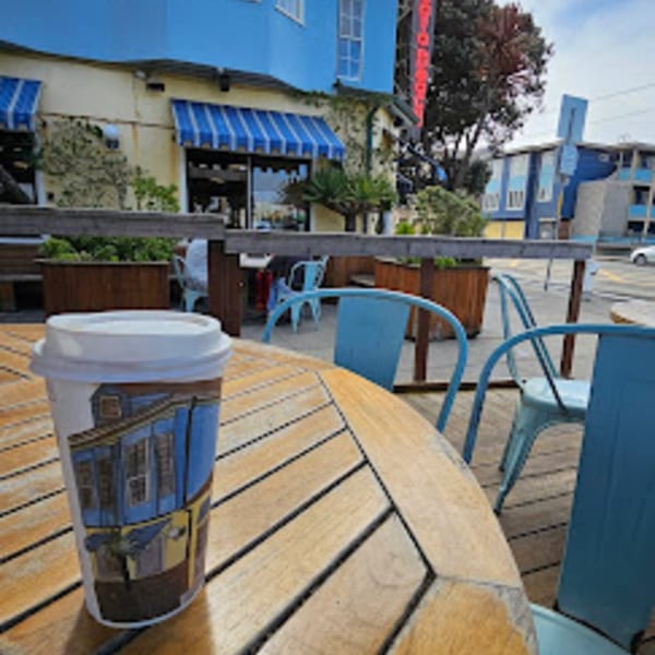A Java Beach coffee cup sits on a wooden outdoor table with blue chairs and cafe in the background.