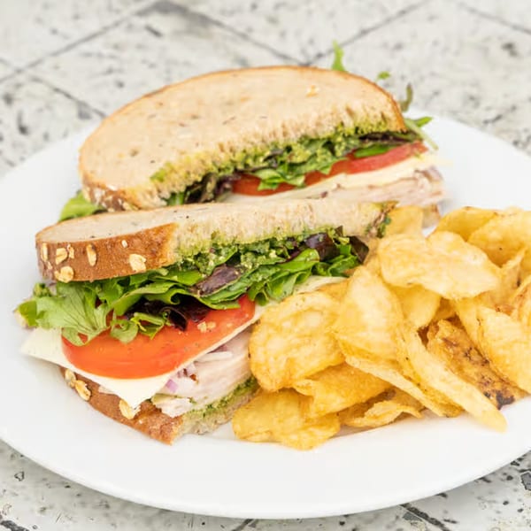 A turkey sandwich with pesto, lettuce, tomato, and cheese, served with potato chips.