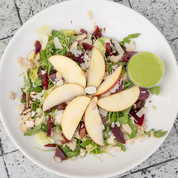 Overhead view of a fresh salad with apple slices, greens, nuts, and a side of green dressing.