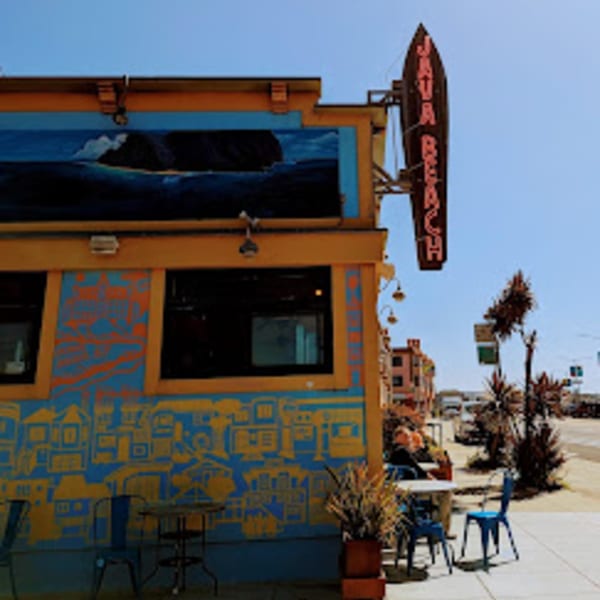 The vibrant blue and yellow exterior of Java Beach cafe with outdoor seating and a neon sign.