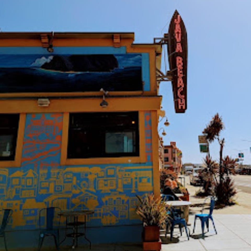 The vibrant blue and yellow exterior of Java Beach cafe with outdoor seating and a neon sign.