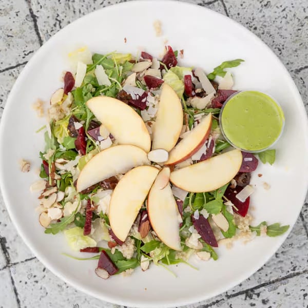Overhead view of a fresh salad with apple slices, greens, nuts, and a side of green dressing.