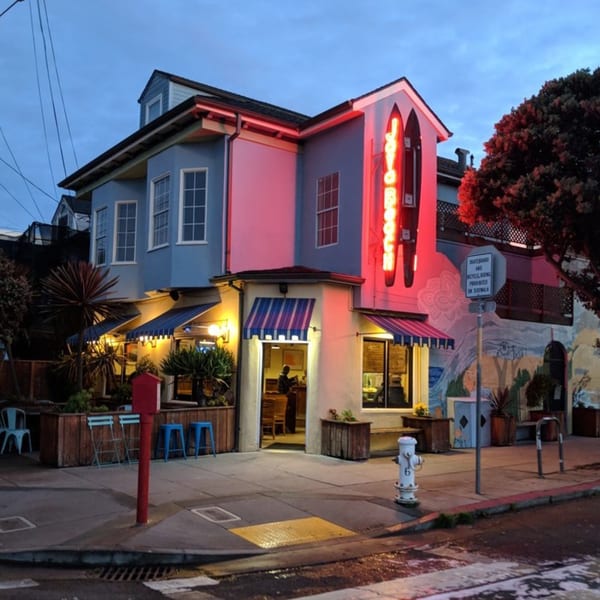 Java Beach cafe exterior at dusk, with blue awnings, red neon sign, and outdoor seating.
